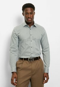 Hellblau gemustertes Hemd mit figurbetontem Schnitt, Bubi-Kragen und Knopfverschluss. Kombiniert mit hellbraunen, plissierten Hosen und einem dunklen Gürtel.