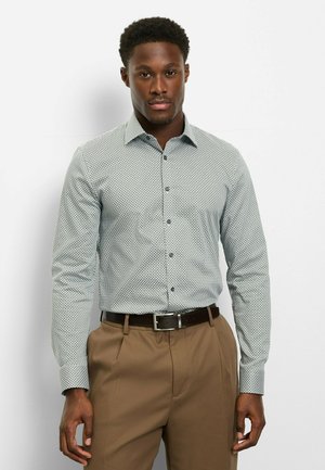 Hellblau gemustertes Hemd mit figurbetontem Schnitt, Bubi-Kragen und Knopfverschluss. Kombiniert mit hellbraunen, plissierten Hosen und einem dunklen Gürtel.