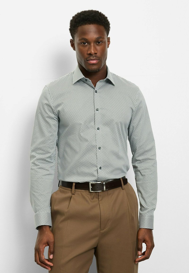 Hellblau gemustertes Hemd mit figurbetontem Schnitt, Bubi-Kragen und Knopfverschluss. Kombiniert mit hellbraunen, plissierten Hosen und einem dunklen Gürtel.