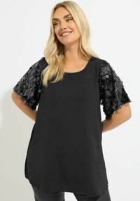 Top nero con corpo testurizzato e maniche corte decorate da paillettes nere, caratterizzato da un collo arrotondato e una vestibilità ampia.
