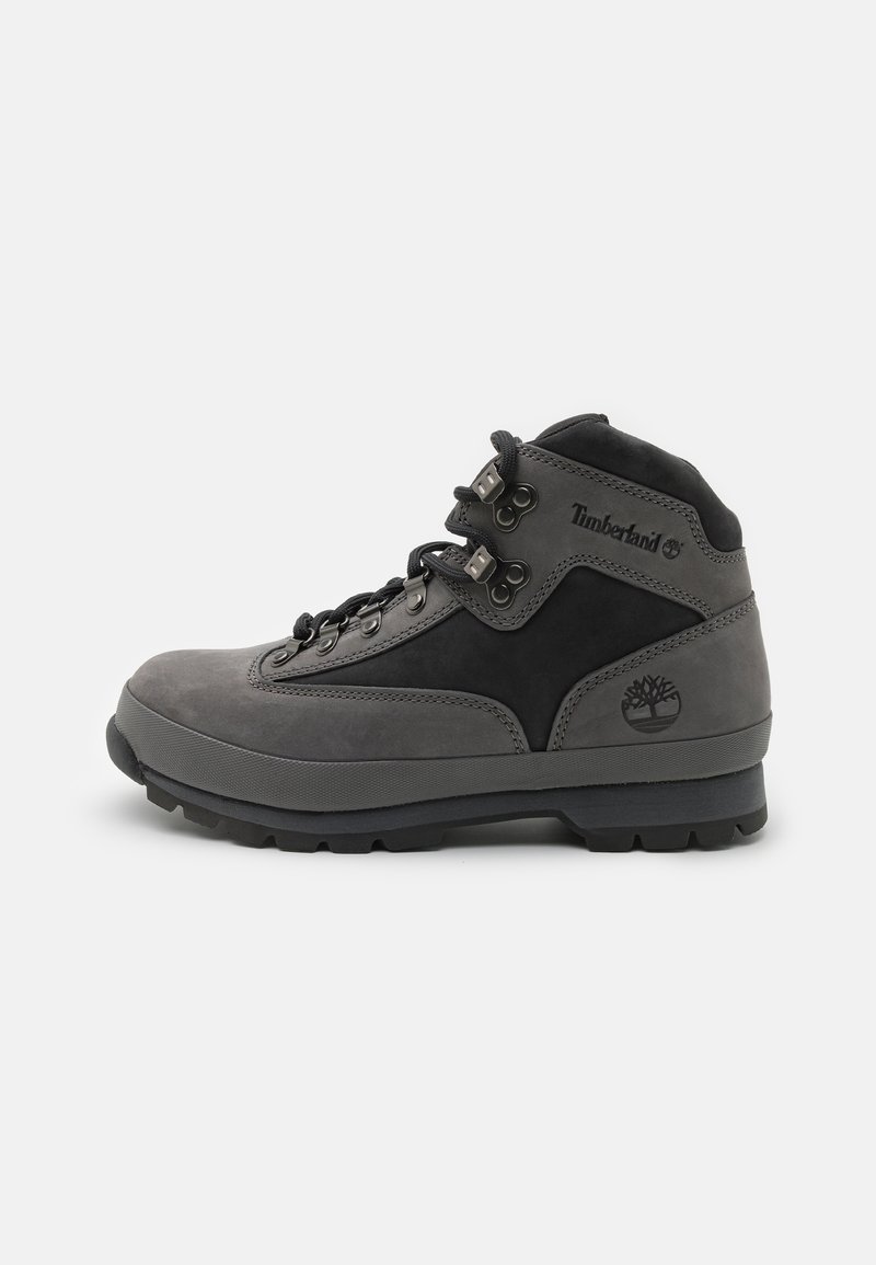 Pilkas žygio batas, pagamintas iš odos ir zomšos medžiagų, su aukštu kulkšnies dizainu, metaliniais akmenukais ir tekstūruotu guminiu padu. Matomas Timberland logotipas.