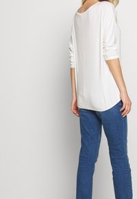 Frau in einem weißen Langarmoberteil mit hochgekrempelten Ärmeln und blauen Jeans, die mit dem Rücken zu einem schlichten hellen Hintergrund steht.