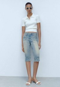 Polo shirt bianco a righe con maniche corte, jeans blu chiaro fino al ginocchio, sandali bianchi, occhiali da sole bianchi, in piedi davanti a uno sfondo neutro.