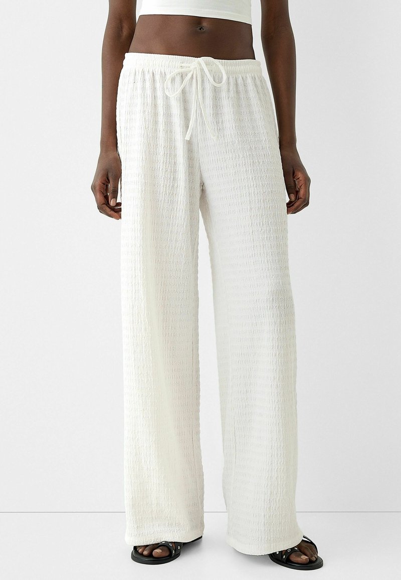 Bílé kalhoty s širokými nohavicemi vyrobené z texturované, zmačkané tkaniny. Mají elastický pas se šňůrkou, vhodné pro volnočasové nošení.