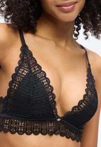 Γυναίκα με μαύρο δαντελένιο σουτιέν με κυματιστές άκρες και λεπτές τιράντες, που εμφανίζει το άνω μέρος του στήθους και μέρος των σγουρών μαλλιών της.