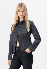 Young woman standing with one hand in pocket wearing dark denim jacket and matching jeans against white background.