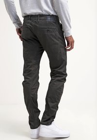 Mörkgrå bomulls-chinos med slim fit, utrustade med bakfickor och en synlig varumärkesetikett på midjebandet, kombinerade med vita sneakers.