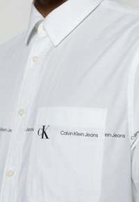 Vit skjorta med knappar som har en strukturerad krage, bröstficka och tryckt logotyp längs fickan och fållen i svart. Mjuk, slät textur.