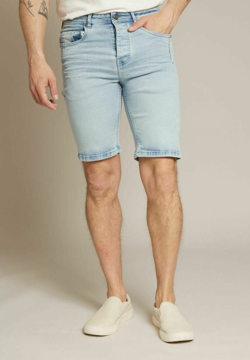 Shorts en denim bleu clair avec un ourlet brut et des poches avant, associés à des chaussures blanches à enfiler. Texture lisse, coupe standard, longueur mi-cuisse.