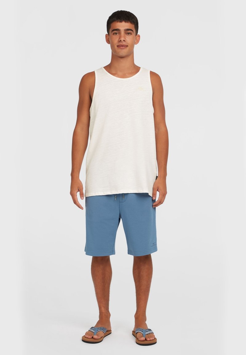 Witte mouwloze tanktop gecombineerd met blauwe shorts, voorzien van een elastische tailleband en trekkoord. Model draagt bruine slippers.
