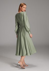 Robe en satin vert avec de longues manches bouffantes, taille cintrée et jupe évasée fluide. Portée avec des talons argentés à lanières.