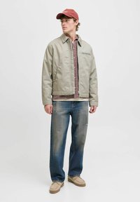 Lys grå jakke med en frontlomme, brunternet skjorte under, parret med blege blå jeans og brune sko. Rød kasket på hovedet.