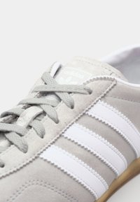 Pelēki zamšādas sporta apavi ar baltām ādas svītrām. Iezīmē apaļu purngalu, teksturētus auklas un gumijas protektoru. Logo uz valnīša.