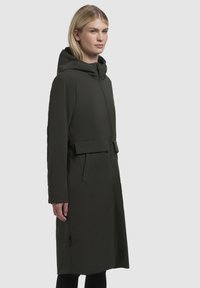 Cappotto lungo verde scuro con cappuccio, dotato di zip, vita con cintura e tasche laterali. Tessuto liscio con design minimalista.