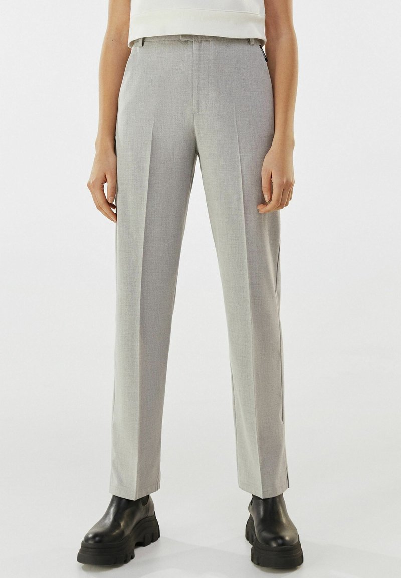 Femme portant un pantalon gris clair sur mesure et des bottes noires épaisses, debout devant un fond blanc uni.