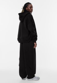 Schwarzer, oversized Hoodie mit Kordelzugkapuze, kombiniert mit lockeren schwarzen Hosen. Graue Turnschuhe mit Netzdetails. Einfaches, minimalistisches Design.