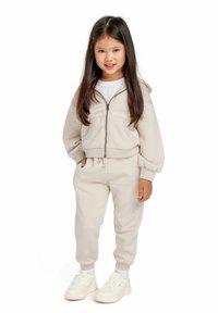 Beige huva zip-up sweatshirt med framfickor och manschetter på ärmarna, parat med matchande joggers. Texturerad bomullstyg och vita sneakers.