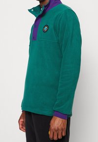 Grön fleecetröja med lila krage, försedd med knappknäppning och en rund logotyp på vänster bröst. Mjuk textur.