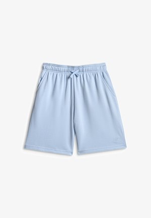 Garçon debout les mains dans les poches, portant un t-shirt blanc oversize, un short bleu clair, des chaussettes blanches et des baskets grises et blanches sur fond gris.
