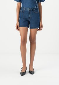 Denim high-waisted shorts in dark blue, featuring a fitted design, five pockets, and a rolled hem, paired with black pointed-toe shoes.