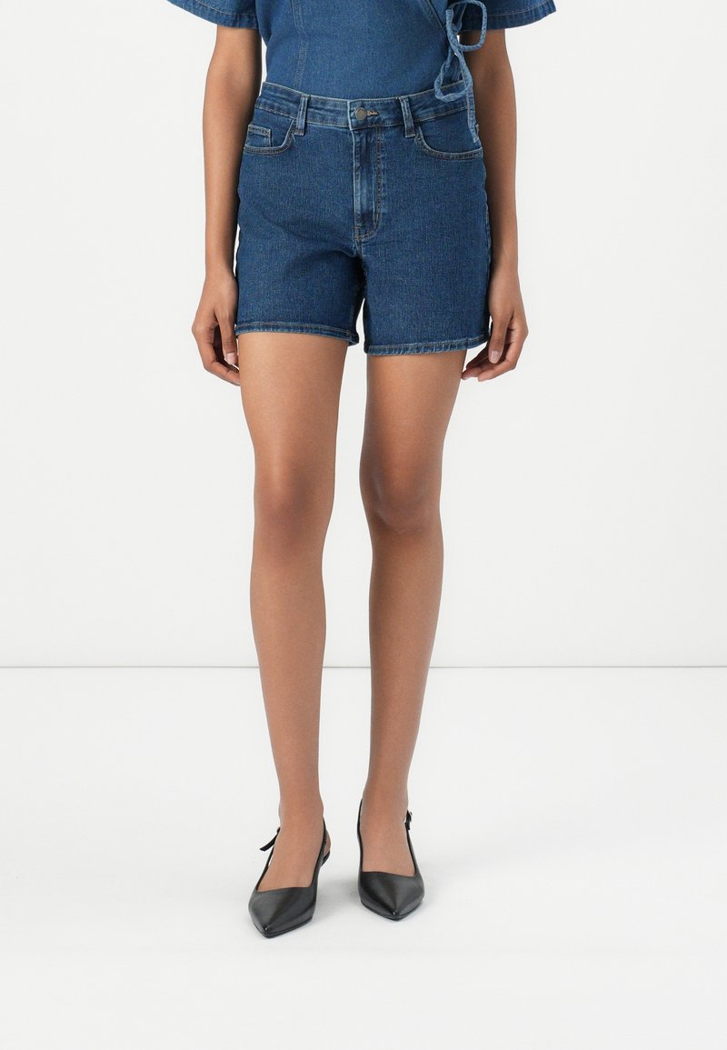 Denim high-waisted shorts in dark blue, featuring a fitted design, five pockets, and a rolled hem, paired with black pointed-toe shoes.