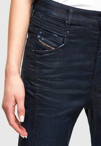 Jeans de mezclilla azul oscuro con un acabado texturizado, que presentan un bolsillo con cremallera, detalles de costura y una etiqueta de logo cerca del bolsillo delantero.