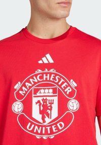 Rotes Baumwoll-T-Shirt mit einem großen weißen Wappen von Manchester United, auf dem ein Teufel einen Dreizack hält, umrahmt von Fußballakzenten und Text.