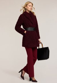 Burgundský kabát z umělé kožešiny s černým opaskem, spárovaný s ladícími burgundskými kalhotami a tmavými podpatky. Drží černou tašku s průhledným držadlem.