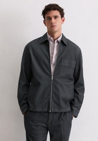 Ciemnoszara kurtka na zamek z kołnierzem i pojedynczą kieszenią na piersi, wykonana z teksturowanej tkaniny, zestawiona z koszulą w paski pod spodem.