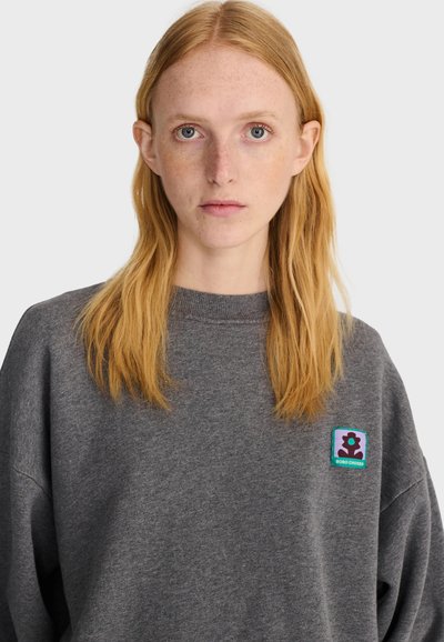 Femme aux longs cheveux roux et aux légères taches de rousseur, portant un pull gris ample avec un écusson coloré sur la poitrine gauche sur un fond uni.