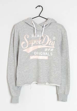 Jasnoszara krótka bluza z kapturem z białymi sznurkami zawieszona na czarnym wieszaku, z tekstem "SuperDry Originals" i japońskimi znakami na przodzie.