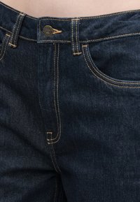 Kroni posnetek pasu temno modrih denim hlač s vidno gumbom in podrobnostmi v rumeni niti.