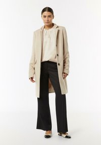 Beiger Wollmantel mit Reverskragen, schwarzen Knöpfen und Seitentaschen, kombiniert mit einer hellen Bluse und schwarzen weiten Hosen.