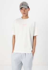 T-shirt blanche à manches courtes avec un logo rose "Hollister", associé à un pantalon rayé bleu clair. La casquette est grise avec un design simple.