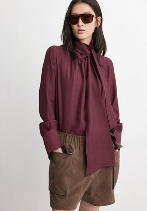 Mujer que lleva gafas de sol con montura carey, una blusa burdeos de manga larga con lazo en el cuello y pantalones cortos de ante marrón con grandes bolsillos.