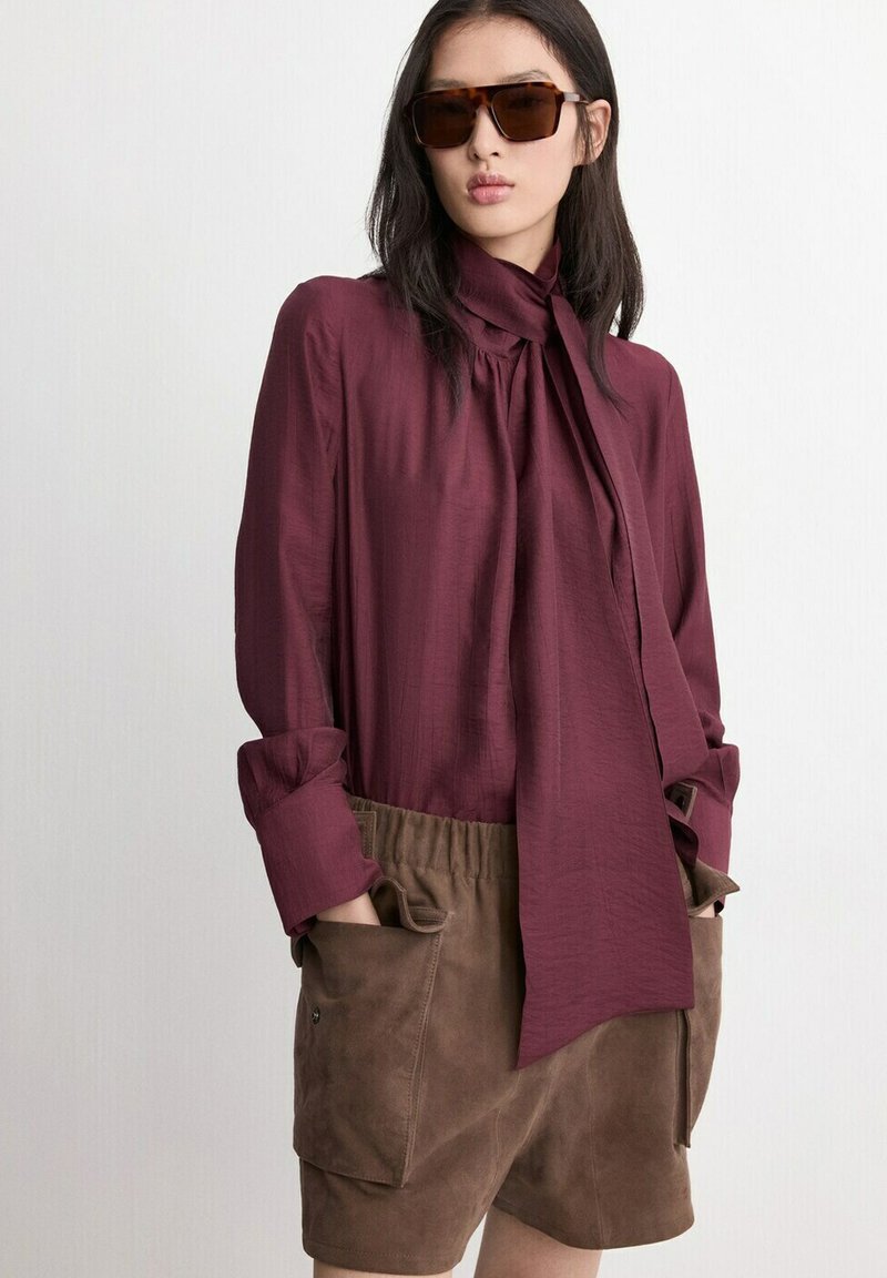Femme portant des lunettes de soleil écaille de tortue, un chemisier bordeaux à manches longues avec un nœud au col, et un short en daim marron avec de grandes poches.
