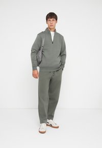 Licht olijfgroen sweat outfit met een pullover met ritssluiting en losse broek, gemaakt van zachte stof. Geaccentueerd door witte sneakers en een grijze tas.