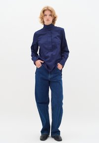 Camicia blu navy a maniche lunghe con colletto alto e pattina frontale. Abbinata a jeans di denim blu larghi e scarpe nere.