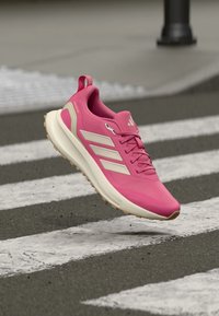 Rosafarbener Sportschuh mit atmungsaktivem Obermaterial, drei weißen Streifen, einer strukturierten Sohle und einem gepolsterten Kragen, der über einem Zebrastreifen positioniert ist.