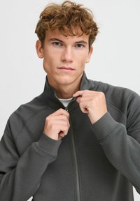 Dark gray zip-up jacket made from smooth fabric, featuring a high collar and ribbed cuffs. The model is holding the zipper halfway closed.
