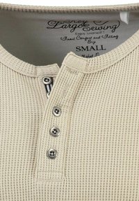 Maglietta a coste beige con scollo rotondo, dotata di tre bottoni metallici sulla patta. L'etichetta indica la taglia small.
