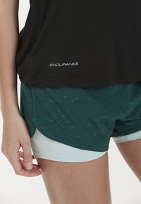 Svart idrottstopp med "ENDURANCE"-logotyp, tillsammans med gröna mesh-shorts som har ett geometriskt utskärningsmönster och ljusblåfoder.