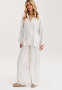 Bílá lehká blůza a kalhoty širokého střihu s složitými paisley vzory, s knoflíkovým zapínáním vpředu a volným střihem. Doplněné o měkkou béžovou obuv.