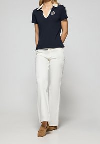 Kvinna klädd i marinblå polo med vit krage och emblem, vita raka byxor och bruna mockaskor, stående med händerna bakom ryggen.