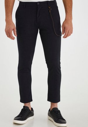Pantalones taper en azul oscuro con una textura suave, presentan un cierre frontal con botón y un acento decorativo de cadena en el lateral.
