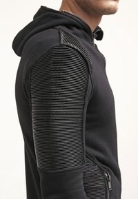 Felpa nera con zip, caratterizzata da accenti neri a costine sulle spalle, tessuto texturizzato sulle maniche e una cappuccio foderato.