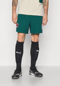 Zielone spodenki sportowe z logo drużyny, czarne skarpetki do kolan z białym logo oraz czarne sneakersy z teksturowaną powierzchnią.