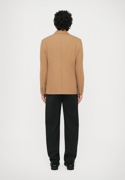 Blazer marrón con un corte entallado, doble abertura y detalles en botones dorados, combinado con pantalones de pierna ancha negros y zapatos negros.