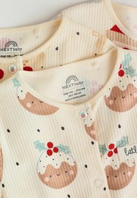 Cremefarvede ribbede babysnapsuits med julepudding mønstre, prikkede accenter og logo-tags; front snap-lukninger forbedrer brugervenligheden.