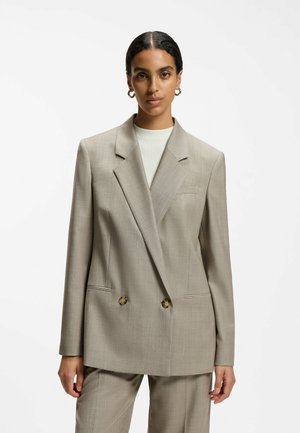 Femme aux cheveux foncés portant un blazer beige croisé et un pantalon assorti, avec un haut blanc côtelé et des boucles d'oreilles créoles dorées.